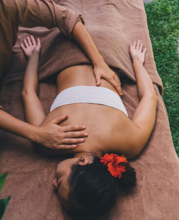 Healthy female with flower in hair lying at massage desk and relaxing during spa procedure with professional, Asian client recreating during beauty therapy at nature feeling calmness and harmony