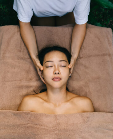 Relaxed Asian woman lysing at massage desk with towels recreating during beauty procedures with professional cosmetologist, young female feeling carefree during body therapy for healthy rejuvenation